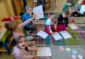 Dzieci siedzą na dywanie i słuchają, jak maja wykonać Kartę Pracy.