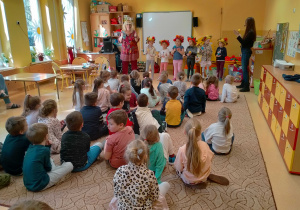 Kilka dziewczynek w kapeluszach przystrojonych owocami i warzywami, śpiewa o nich piosenkę.Reszta dzieci siedzi na dywanie.